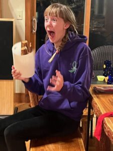 Amy holding a piece of paper and with her mouth open in surprised excitement 