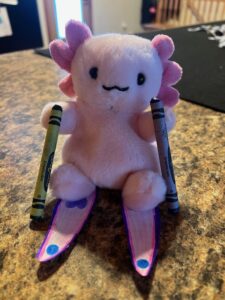 Tiny pink axolotl stuffed animal holding crayons as ski poles and standing on paper skis