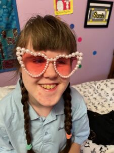 Sarah smiling and wearing pink heart-shaped sunglasses with fake pearls framing each eye piece.