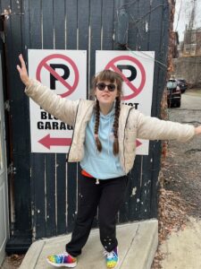 Sarah wearing a white coat, light blue shirt, black sweatpants, rainbow shoes, and sunglasses. Her hair is in two braids that hang in front of her chest. She is standing in front of two No Parking signs with her arms stretched out to either side of her.